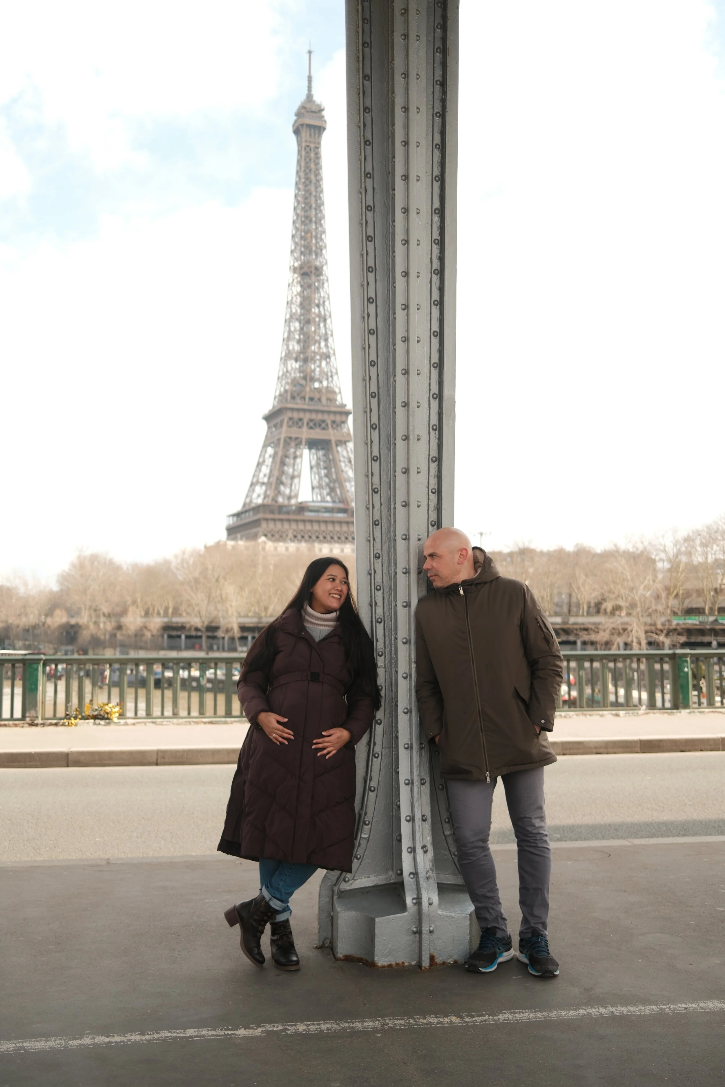 A man and woman standing on a bridge with the Eiffel Tower in the background, leaning against a large metal support column, smiling at each other.