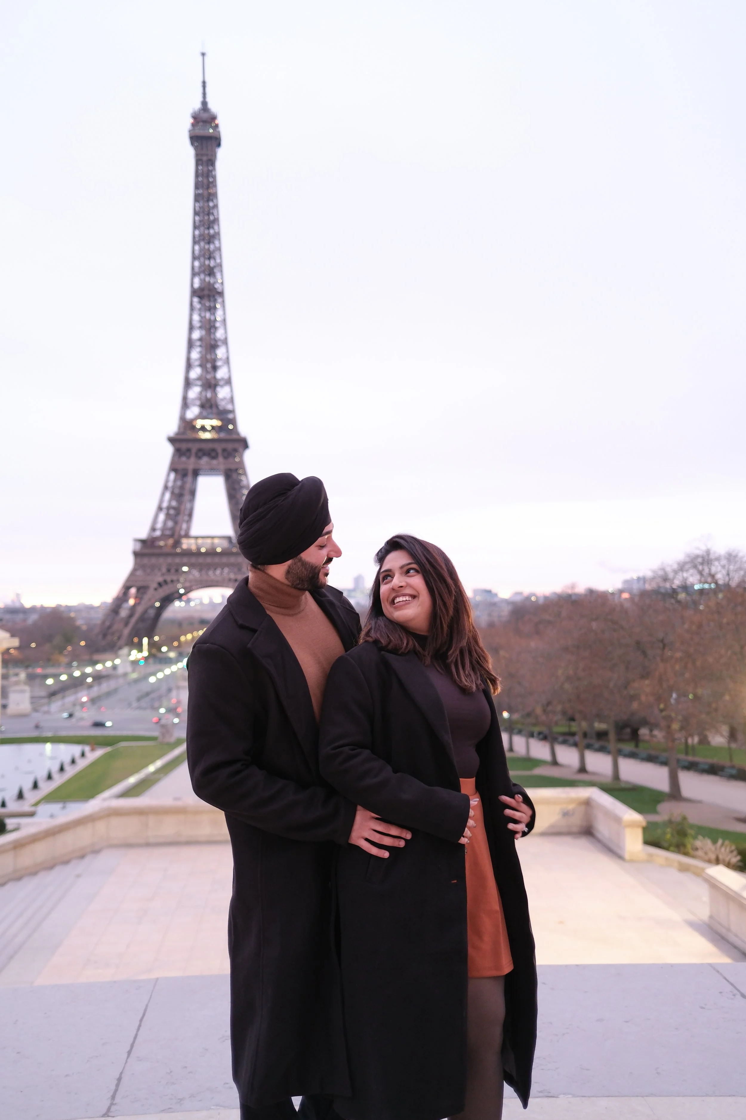A couple standing outdoors near the Eiffel Tower in Paris, France, during the evening, smiling and looking at each other.