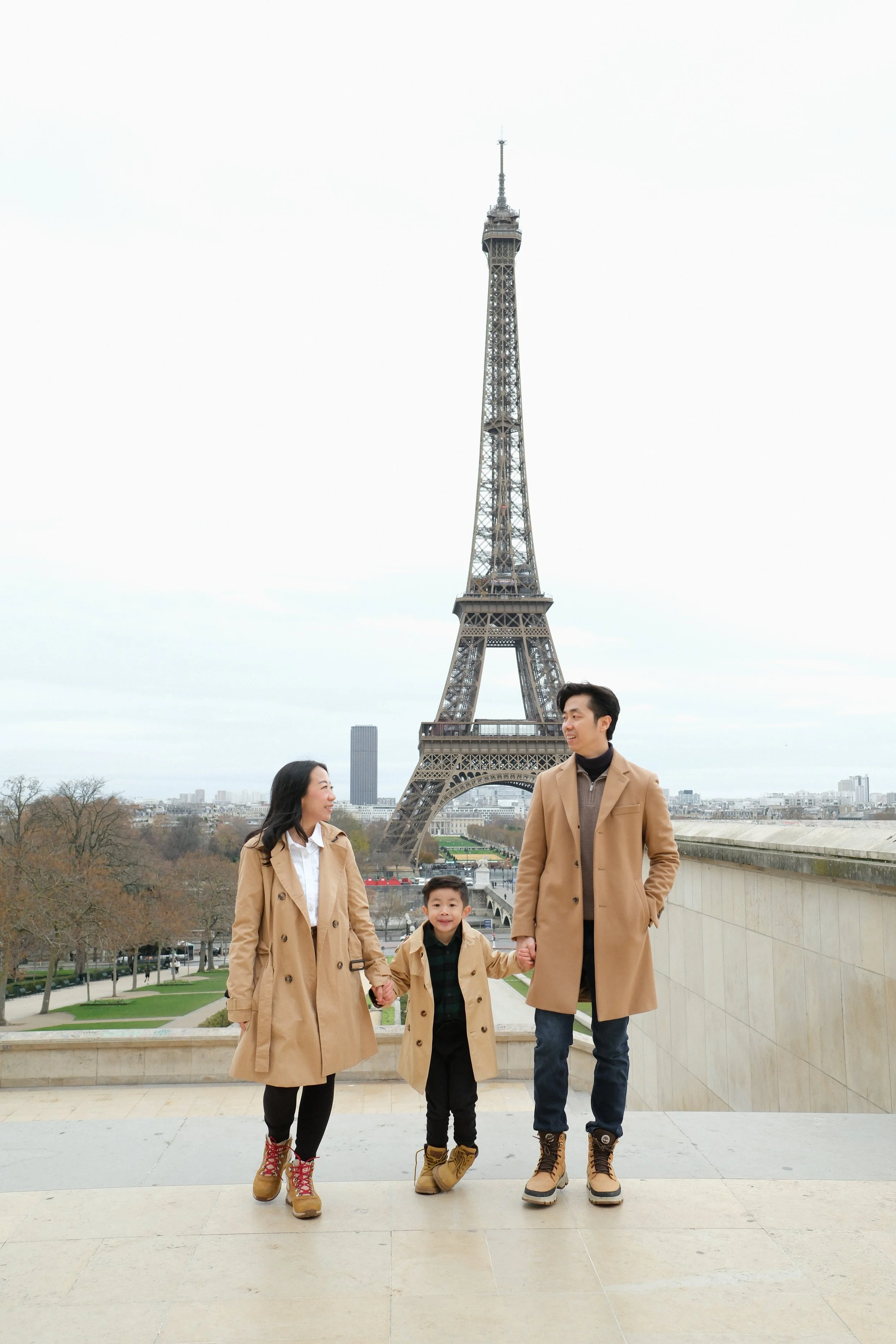 A family of three, all wearing beige coats and boots, holding hands and standing in front of the Eiffel Tower in Paris, France.