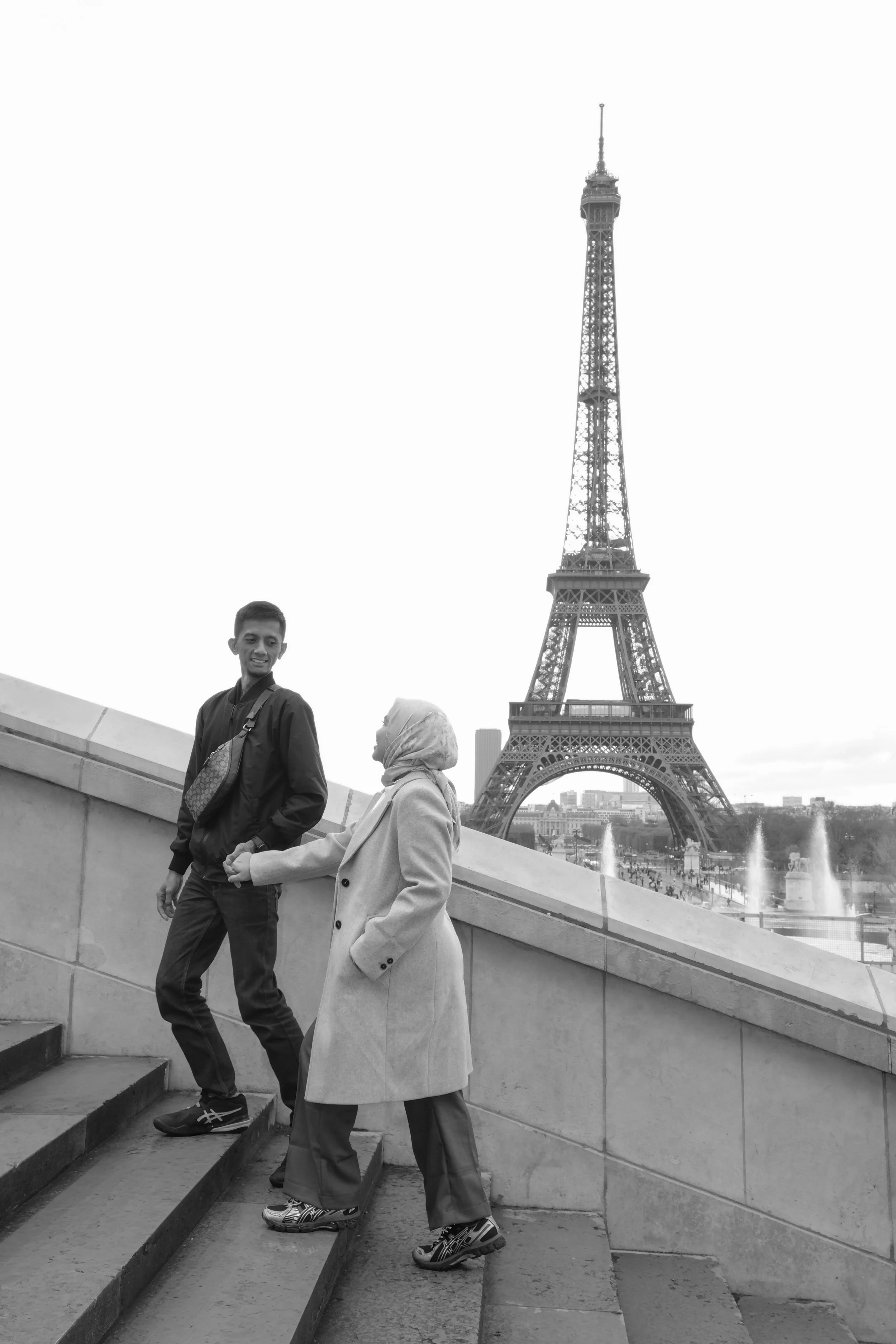 A young man and an elderly woman holding hands and walking up stairs with the Eiffel Tower in the background.