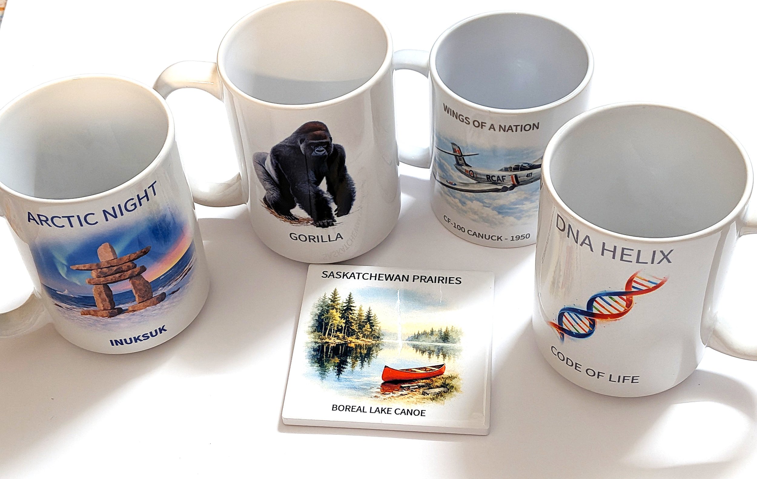 Four white ceramic mugs with different travel or nature-themed images and text, and a tile with a lakeside scene and text. The mugs show Arctic night, a gorilla, wings of a nation jet, and DNA helix respectively, with accompanying descriptive words.