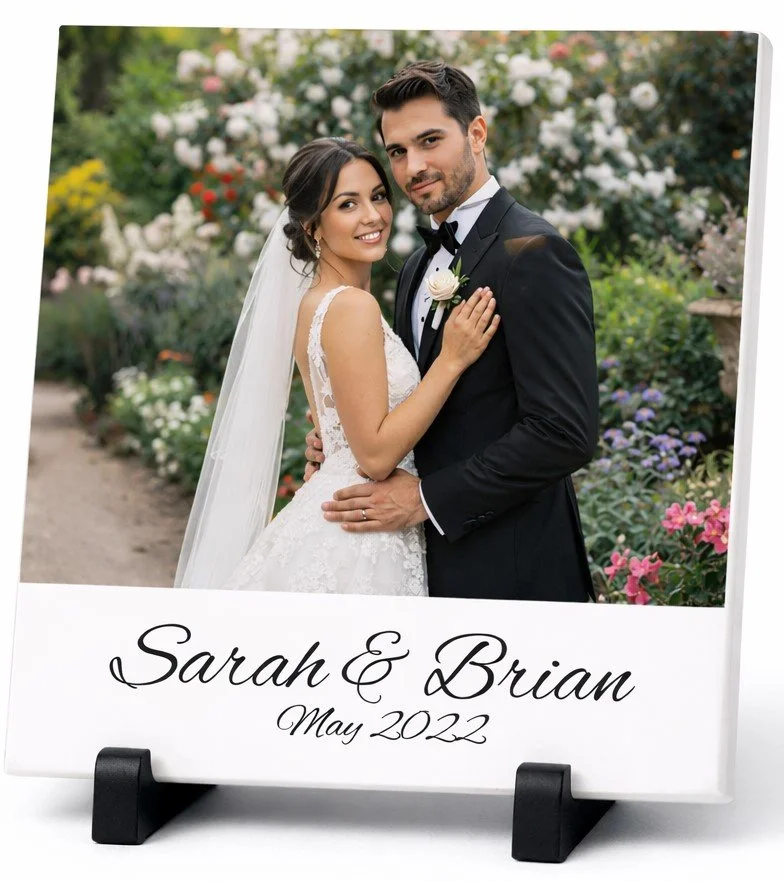 Photograph of a bride and groom in wedding attire standing outdoors in a garden, smiling and embracing each other. The bride wears a white lace wedding dress and veil, and the groom wears a black tuxedo with a bow tie and boutonniere. The image is printed on a personalized display with the text "Sarah & Brian May 2022" at the bottom.