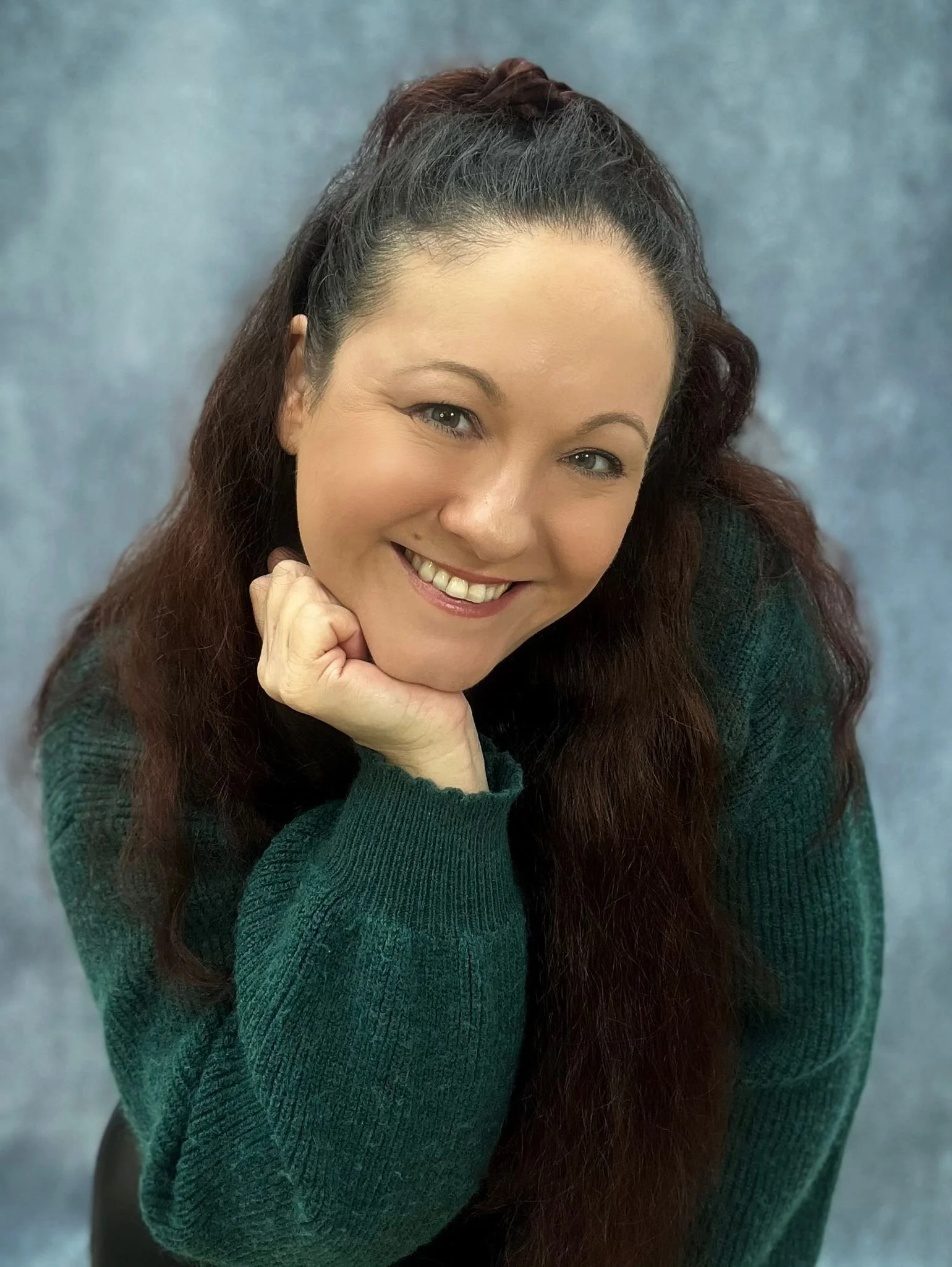 A woman with long, wavy brown hair and light skin is smiling and resting her chin on her hand. She is wearing a green sweater and looking directly at the camera.