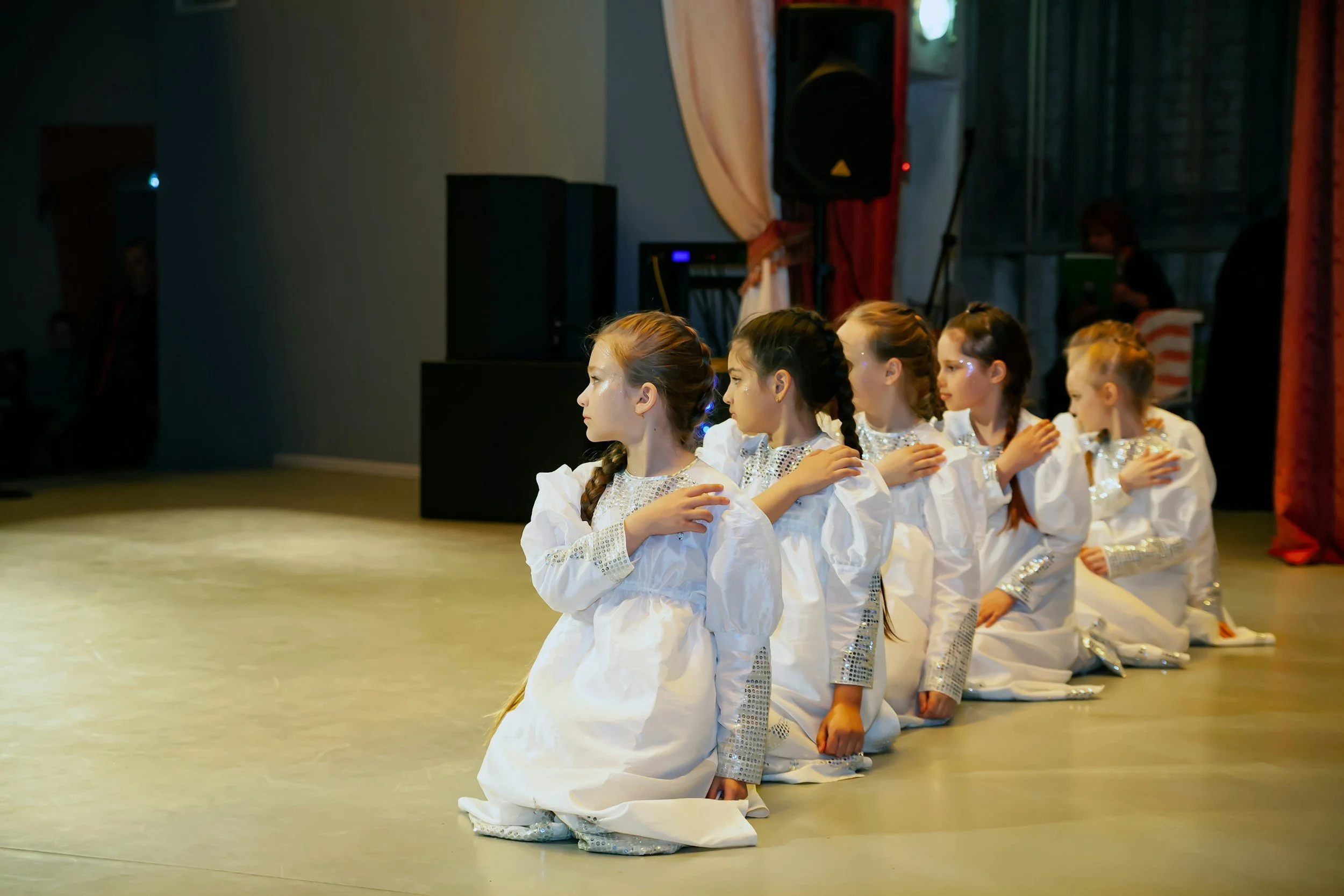 A group of six young girls dressed in white costumes kneeling in a line on a stage, performing a dance or routine with their hands on their chests.