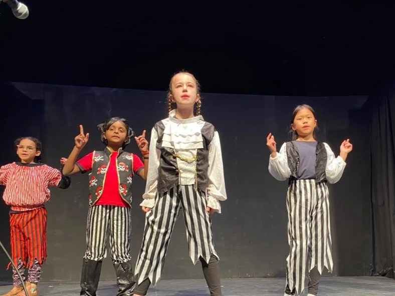Four young girls performing a stage play, dressed in vintage-style costumes, standing on stage with serious expressions.