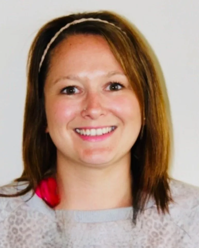 A woman with shoulder-length brown hair, wearing a pink headband and a light-colored patterned shirt, smiling at the camera.