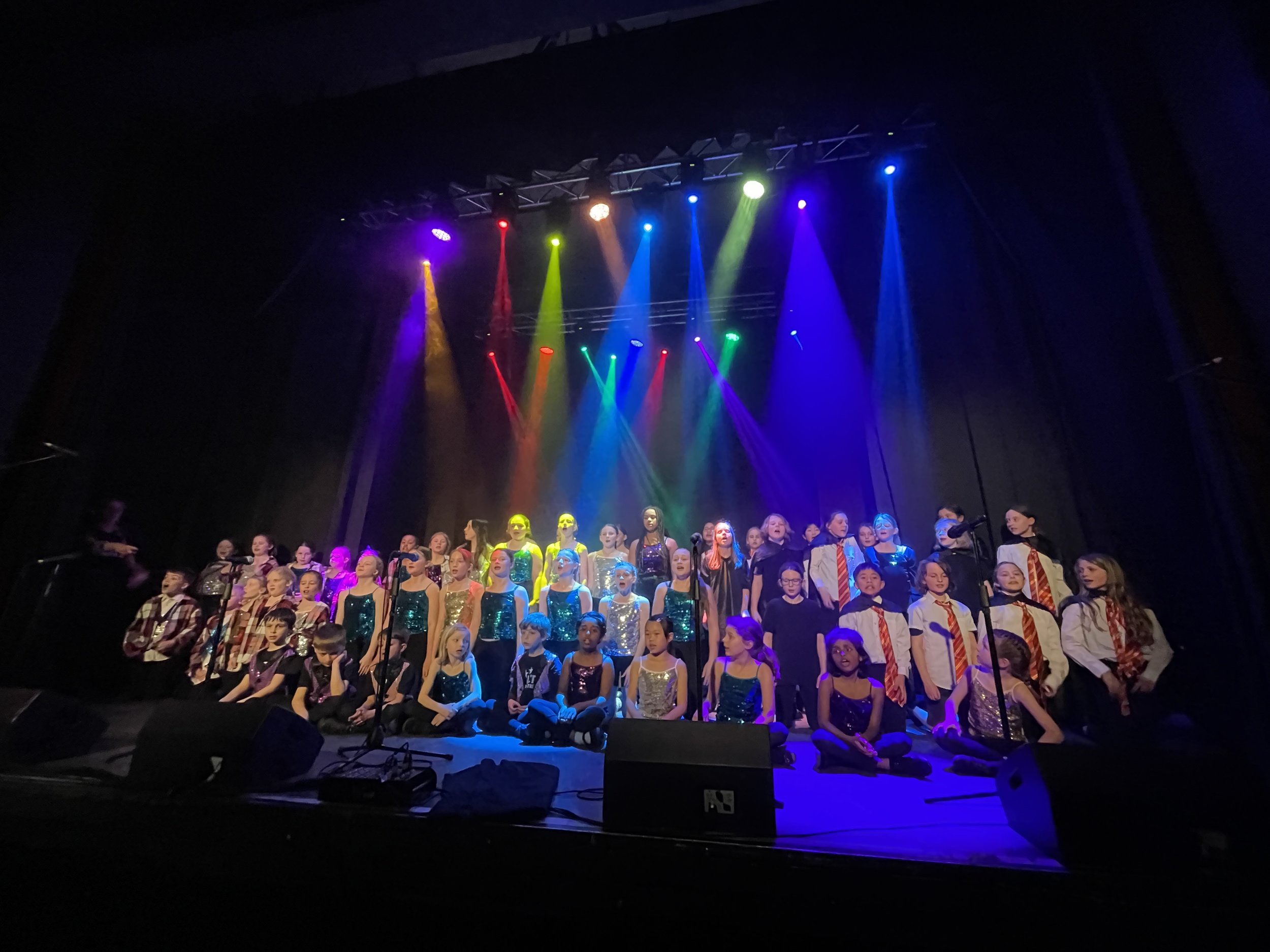 Children performing on stage with colorful spotlights, some are standing and others are sitting, wearing costumes, in a school concert or recital.