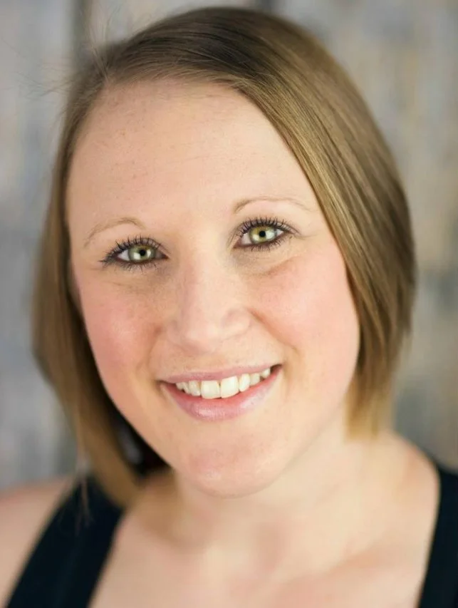 Close-up of a woman with shoulder-length light brown hair, fair skin, green eyes, and a warm smile, wearing a dark top, against a blurred background.