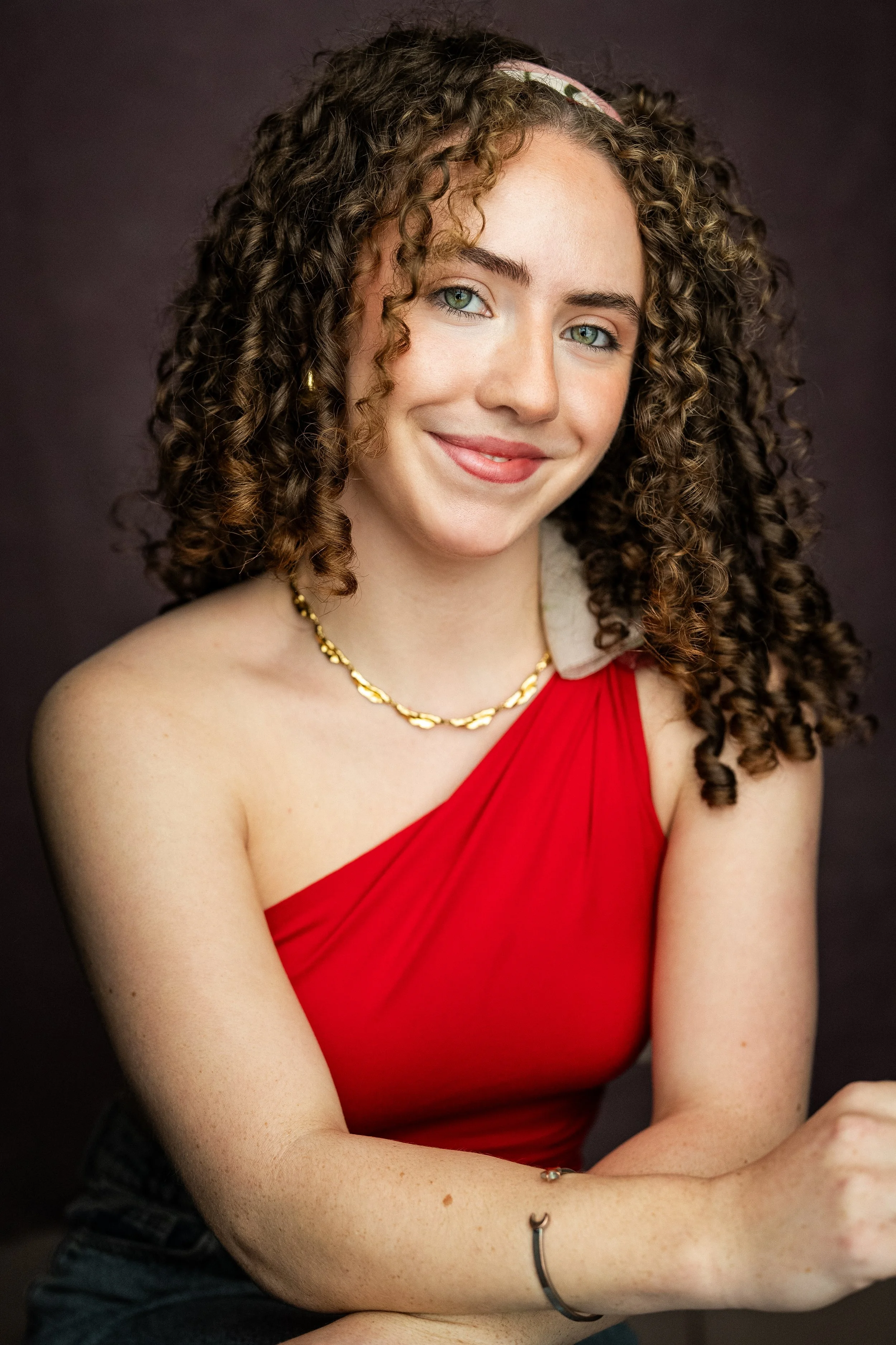 A young woman with curly brown hair, light skin, wearing a red sleeveless top and a gold necklace, smiling at the camera.