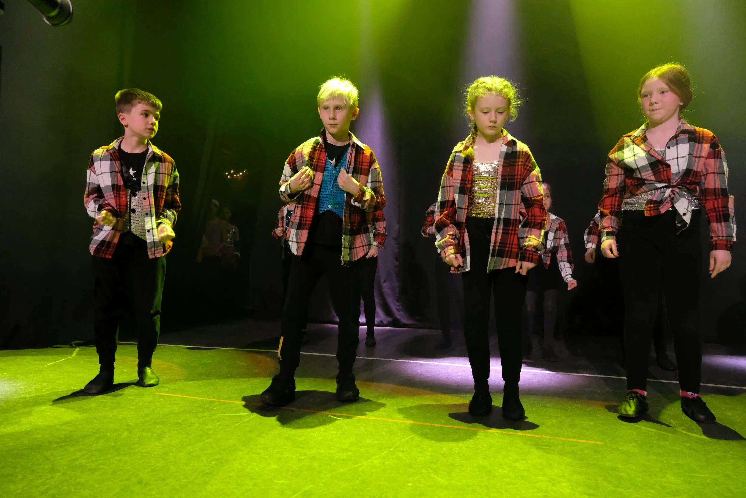 A group of young children performing on stage, wearing plaid shirts and black pants, under green and purple stage lighting.