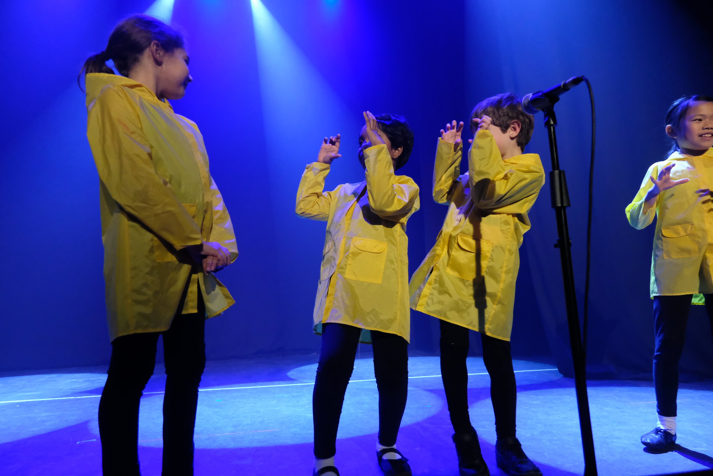 Children wearing yellow raincoats performing on stage with a microphone and blue lighting.