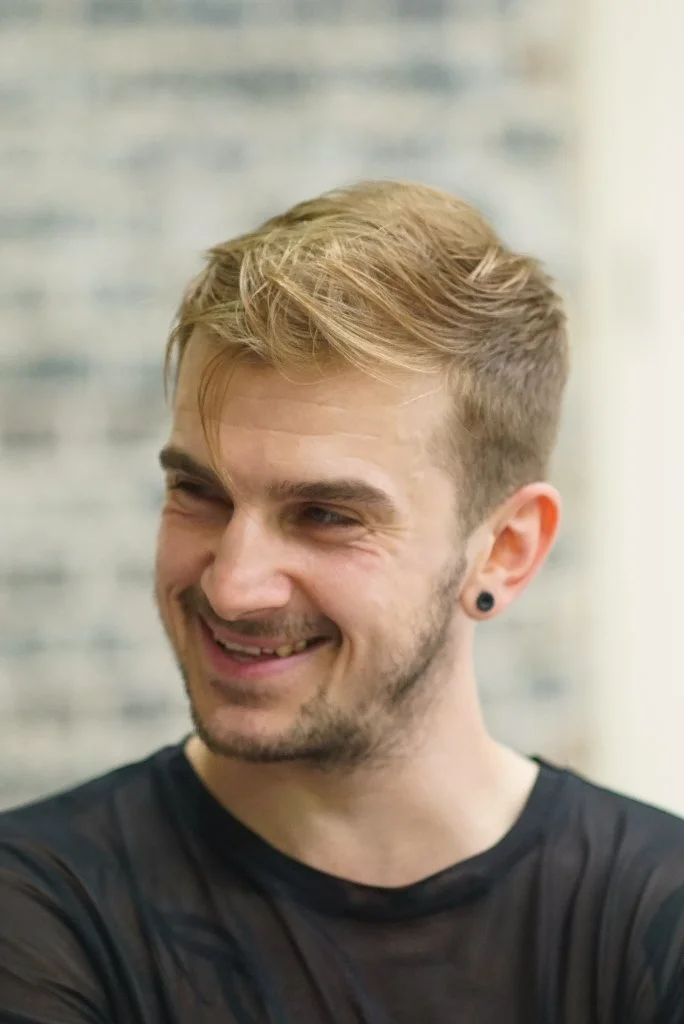 A young man with blonde hair, a light beard, and an earring smiling and looking to the side.