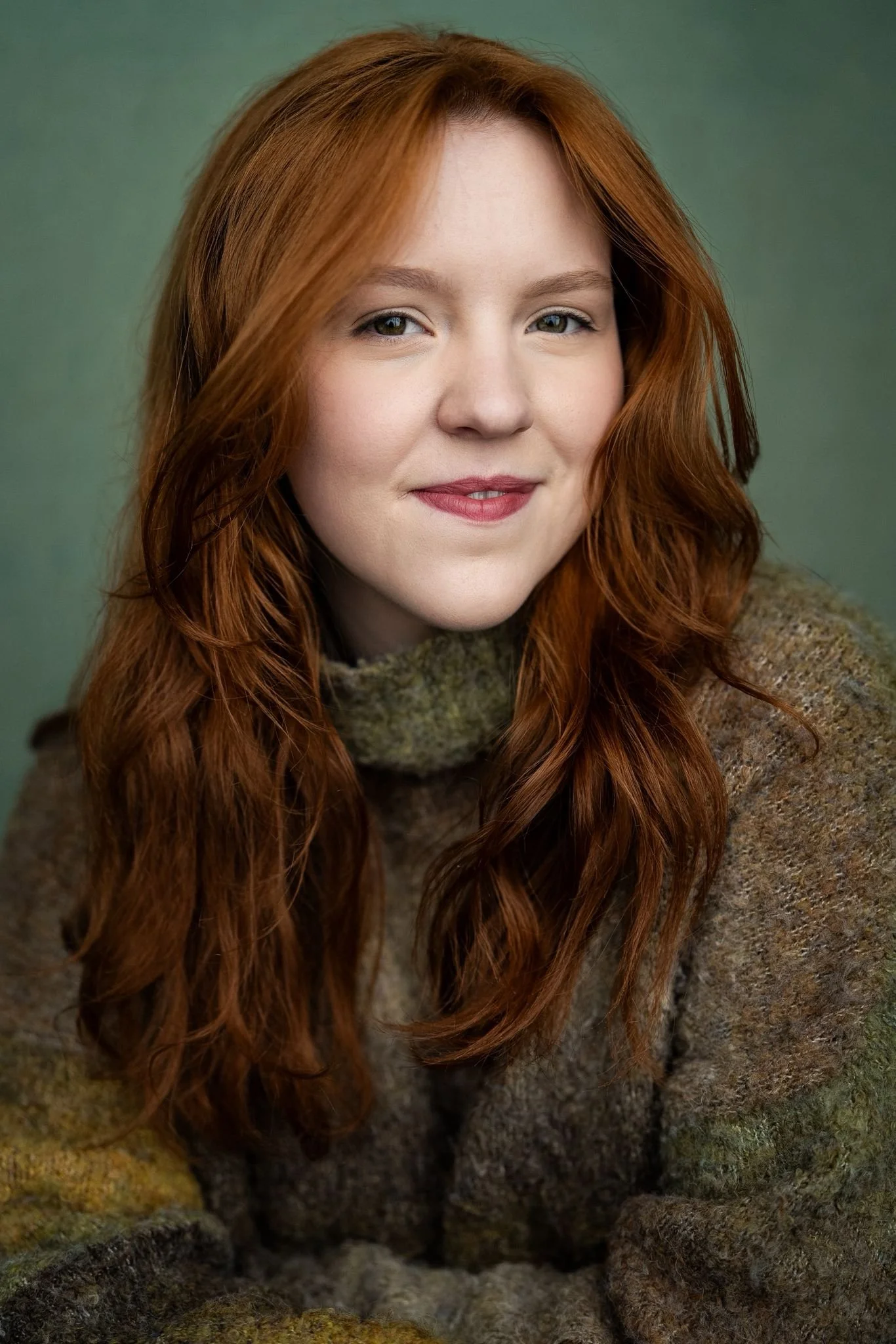 A young woman with long, wavy red hair and fair skin, smiling slightly, wearing a cozy, multi-colored knit sweater, against a muted green background.