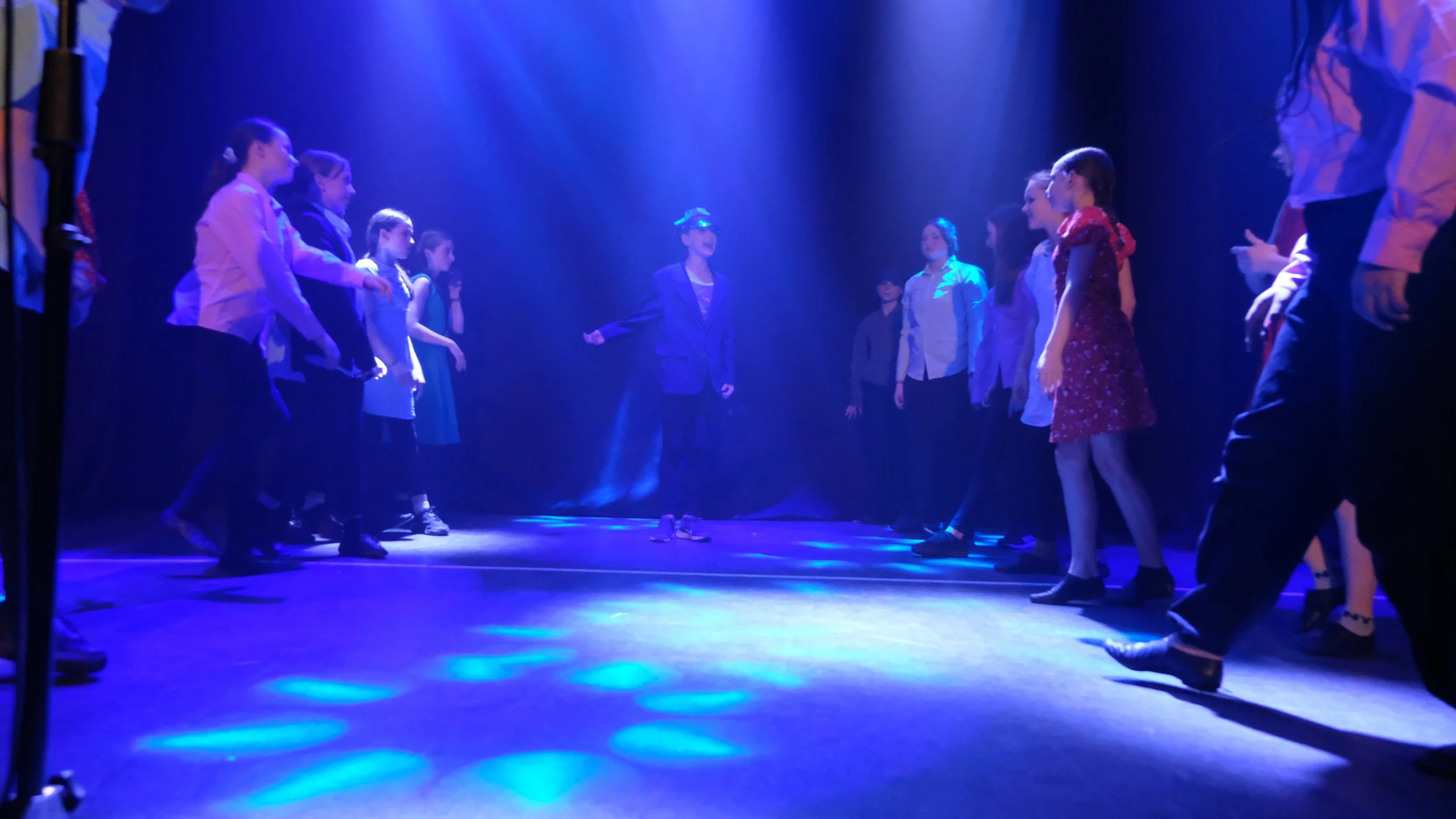Group of children standing on a stage under blue lights, likely preparing for a performance or rehearsal.