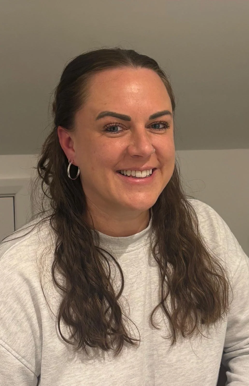 A smiling woman with long brown wavy hair, wearing hoop earrings and a light-colored sweatshirt, sitting indoors in front of a plain wall.