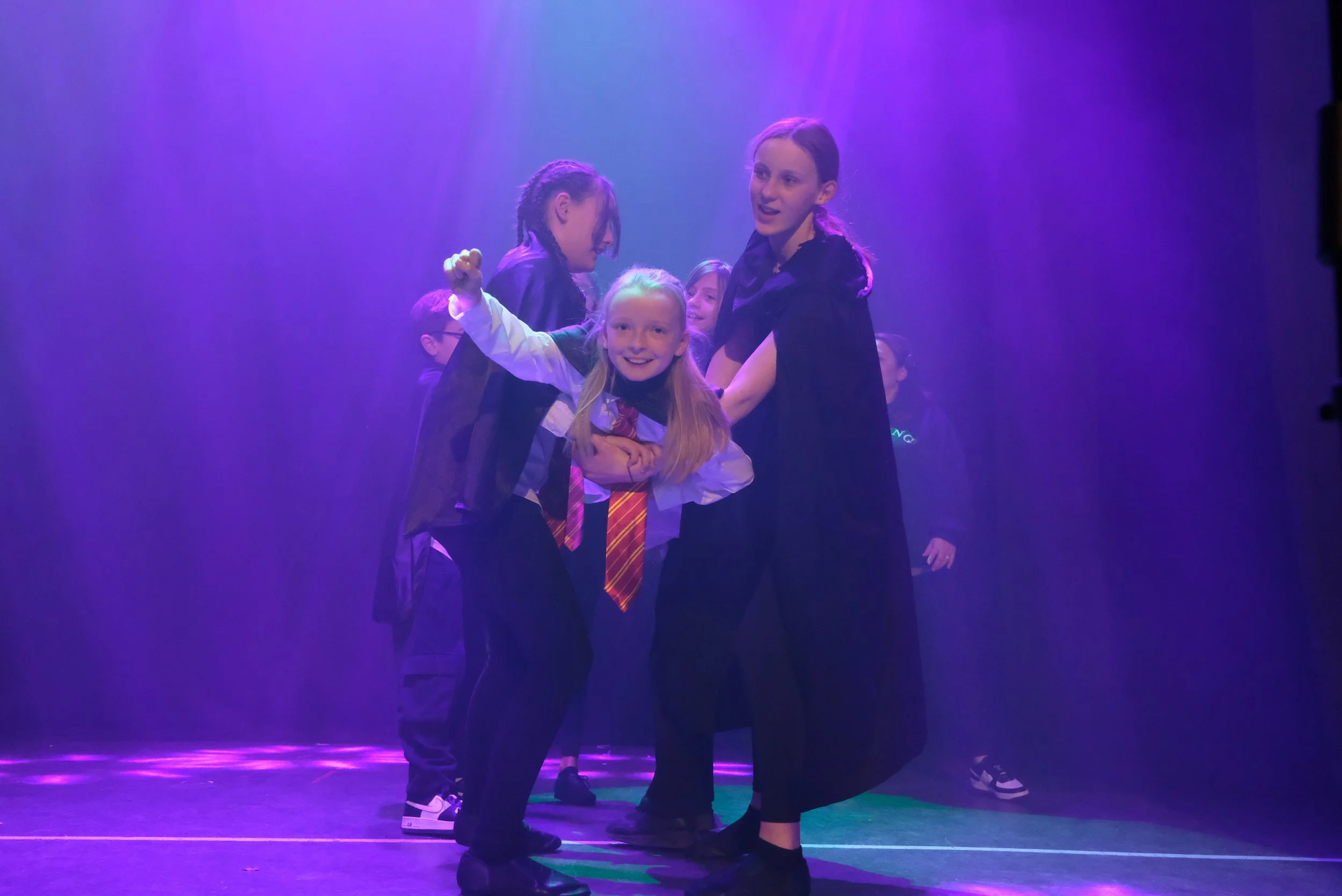 Children dressed as Harry Potter characters on stage, with purple lighting, participating in a play or school performance.