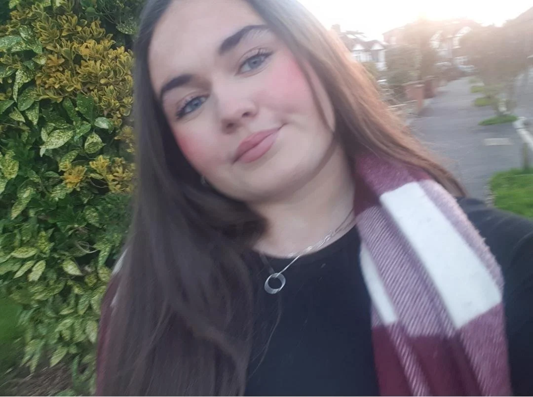 A young woman with long dark hair, wearing a black top and a maroon and white scarf, taking a selfie outdoors in front of bushes and a suburban street at sunset.