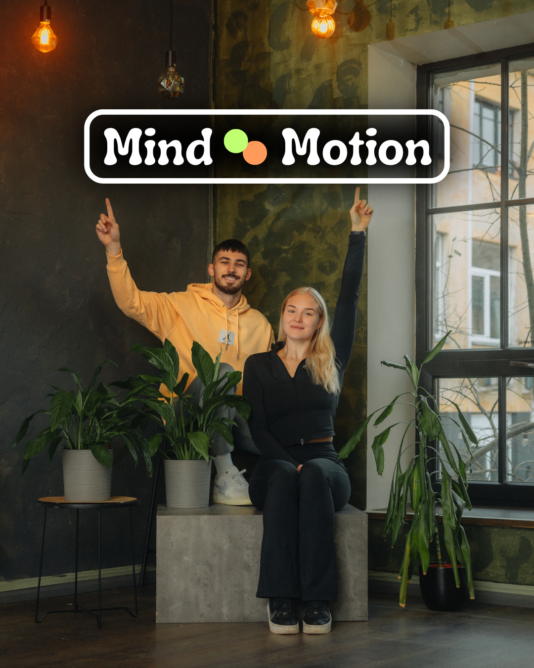 Mind & Motion stuudio asutajad Reiko ja Triin istuvad stuudiotaustal roheliste taimede vahel, käed üles tõstetud. Stuudiokeskkond õdusate lampidega, Tartu Aparaaditehase hoone sees. Mind & Motion logo ülal fotol.