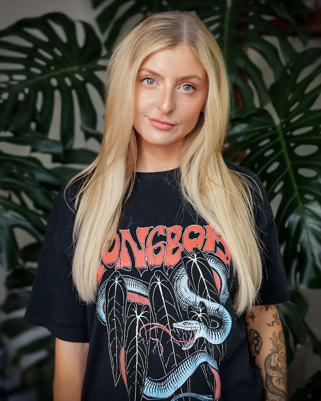 A young woman with long blonde hair, blue eyes, and tattoos on her arm, wearing a black band T-shirt with a snake and tree design, standing in front of large green leaves.