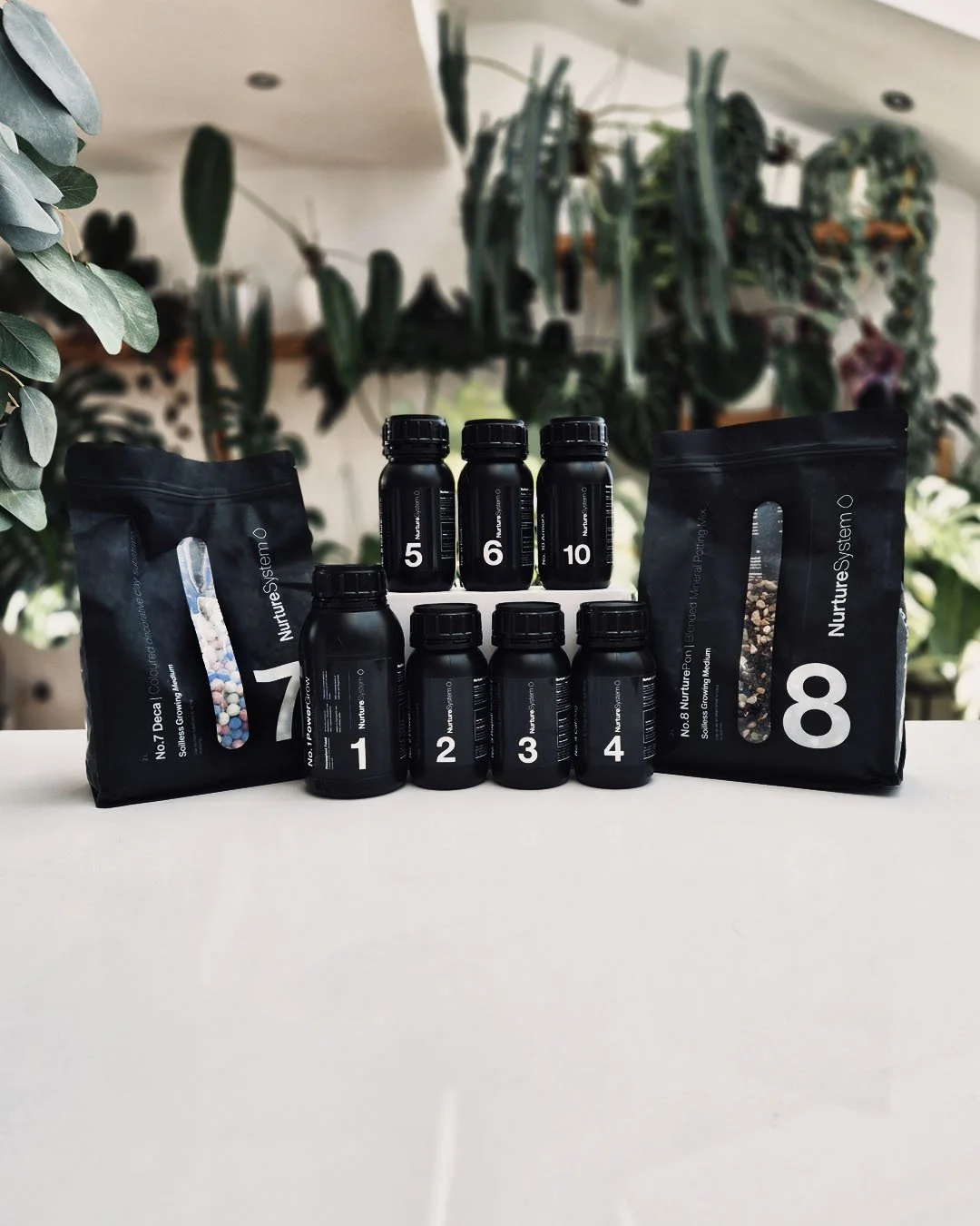 Assorted bottles and packages of plant nutrients or supplements on a white surface with green plants and foliage in the background.