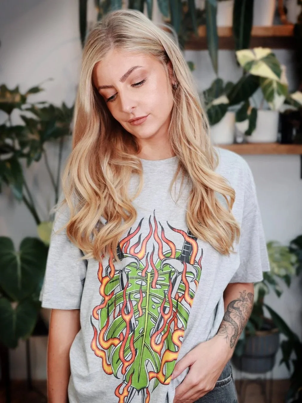 A young woman with long, wavy blonde hair standing indoors, looking down with her eyes closed. She wears a gray t-shirt with a colorful graphic of a stylized green leaf and red-orange flames, and has a tattoo on her left forearm. There are potted plants and shelves in the background.