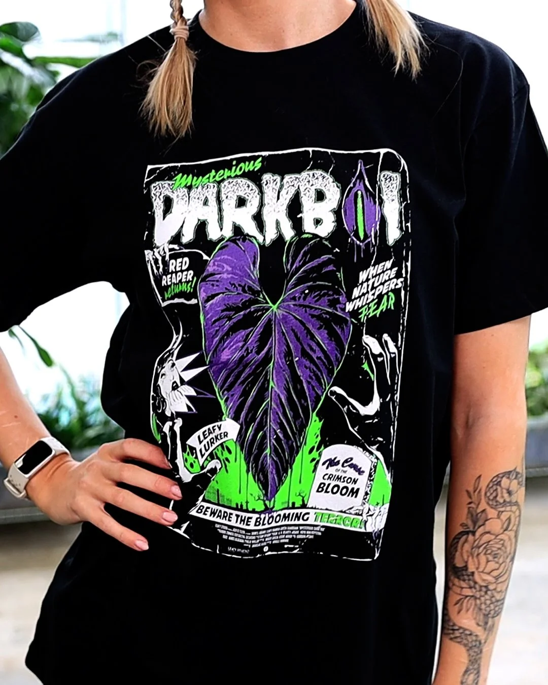 A person wearing a black T-shirt with a graphic design of a purple and green plant, accompanied by text about a mysterious dark plant and warnings about blooming terrarium, in an outdoor setting.