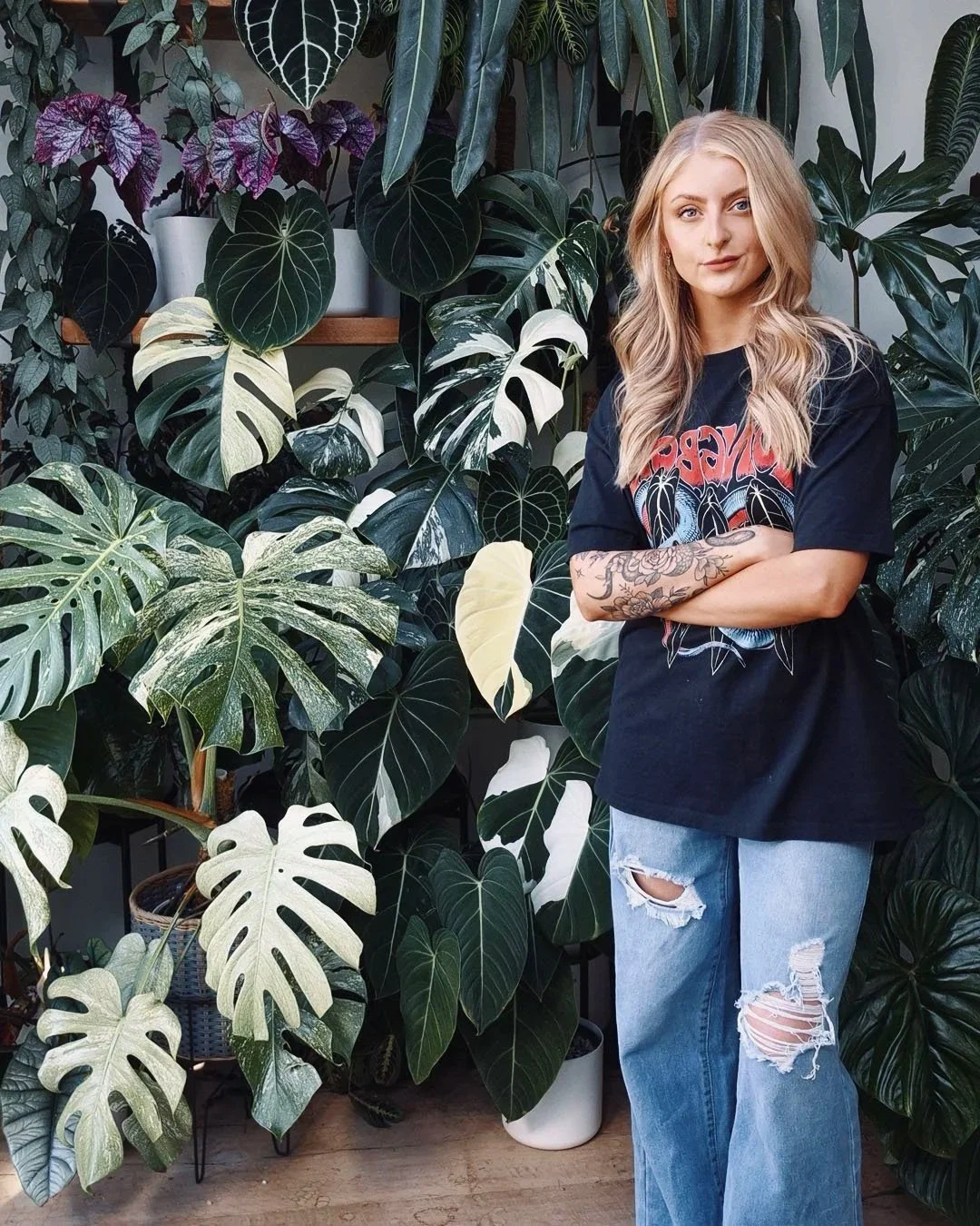 A young woman with long blonde hair standing in front of a large collection of tropical green and variegated houseplants, wearing a black band T-shirt, ripped jeans, and tattoos on her right arm.