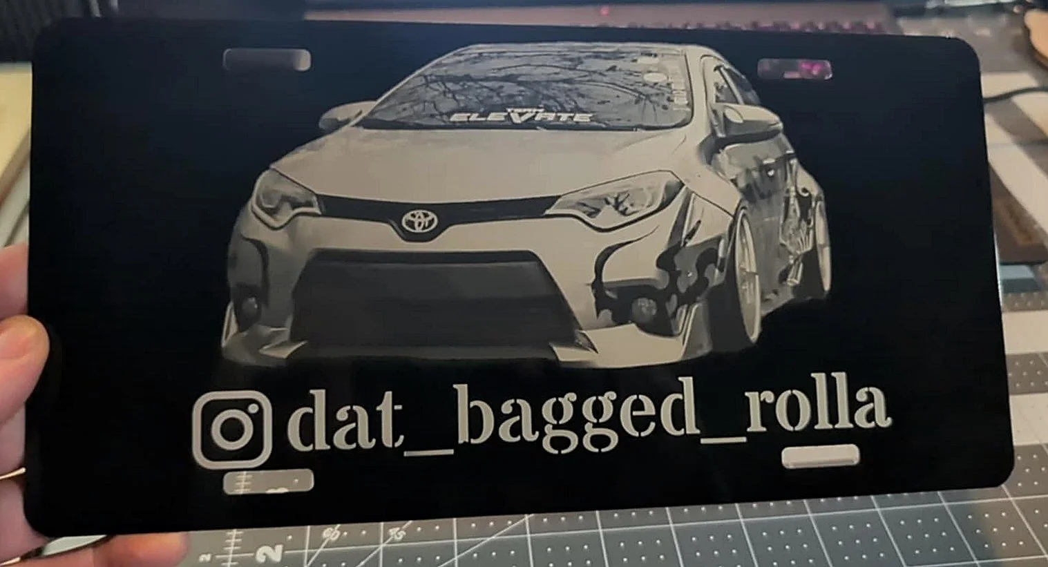 Black license plate engraved with a photo of a bagged Toyota Corolla and the owner's Instagram handle, @dat_bagged_rolla