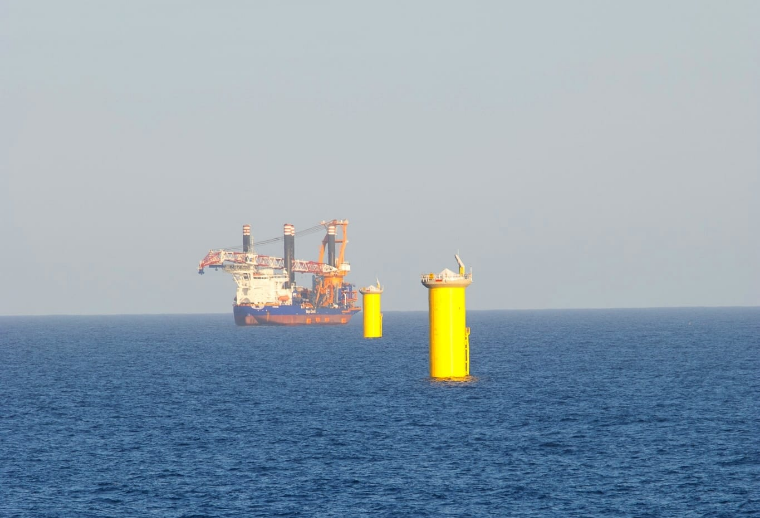 An offshore oil platform in the distance on the ocean, with three large yellow buoys or pillars floating in the water nearby.