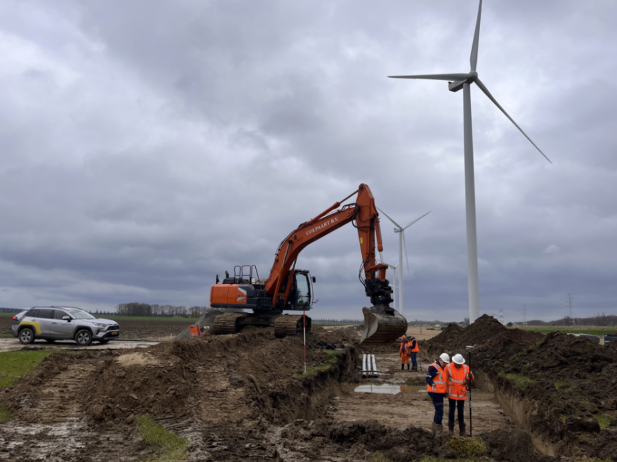 Construction workers in orange safety vests and white helmets working on wind turbine installation with excavator digging trenches, under cloudy sky.