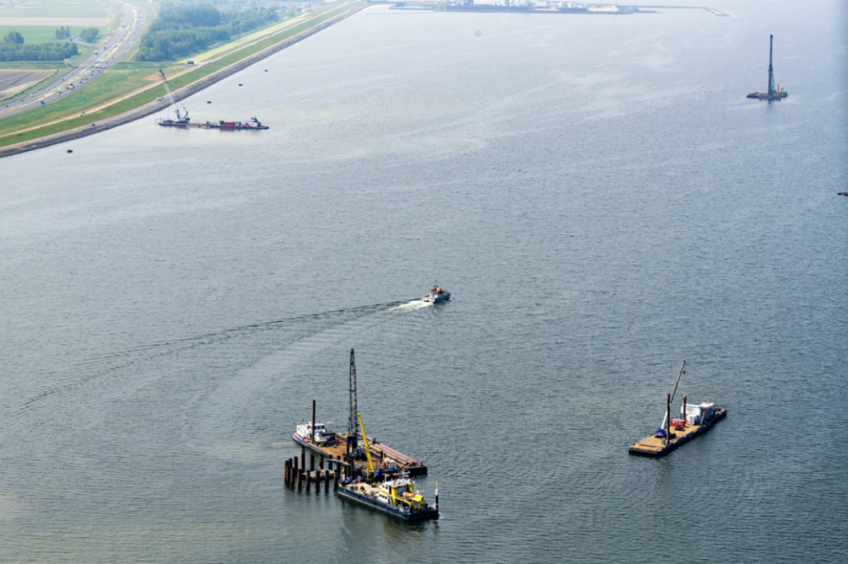 Boats, including a small motorboat, on a large body of water near a shoreline with construction equipment and barges.