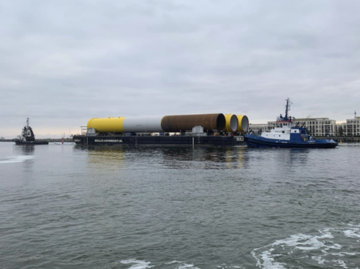 Barge carrying large rocket sections being guided by two tugboats in a harbor, with modern buildings and an overcast sky in the background.