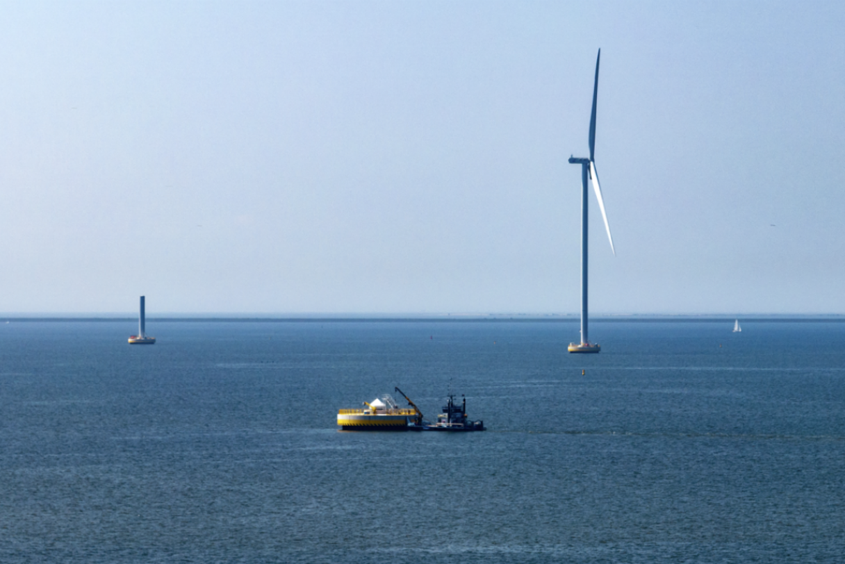 A quote of three wind turbines in a body of water, with a barge in the foreground and small sailboats in the distance.