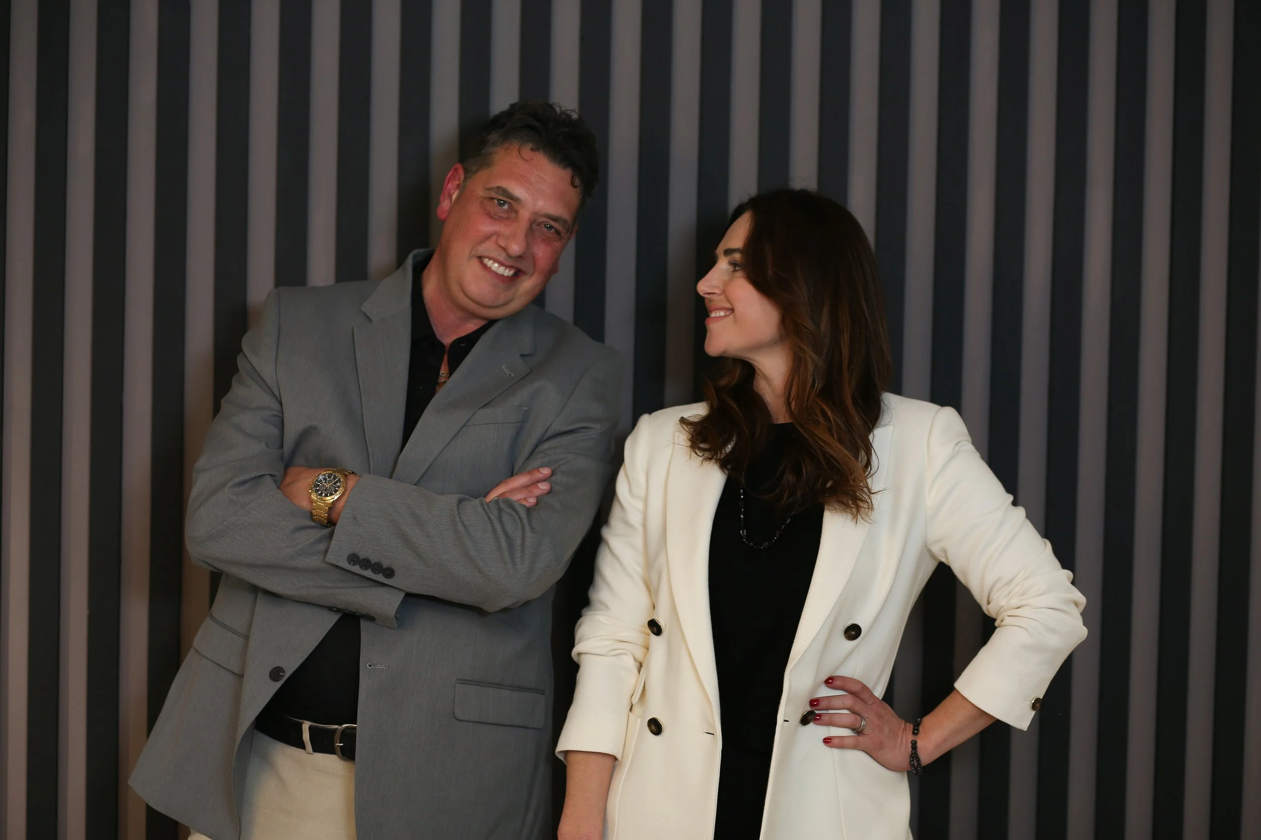 Elliott King and Aleksandra King — husband-and-wife duo, co-authors of Marketing Wins and experts in GEO / AI SEO / video SEO — standing close together, smiling at each other before a black-and-grey vertical striped backdrop.