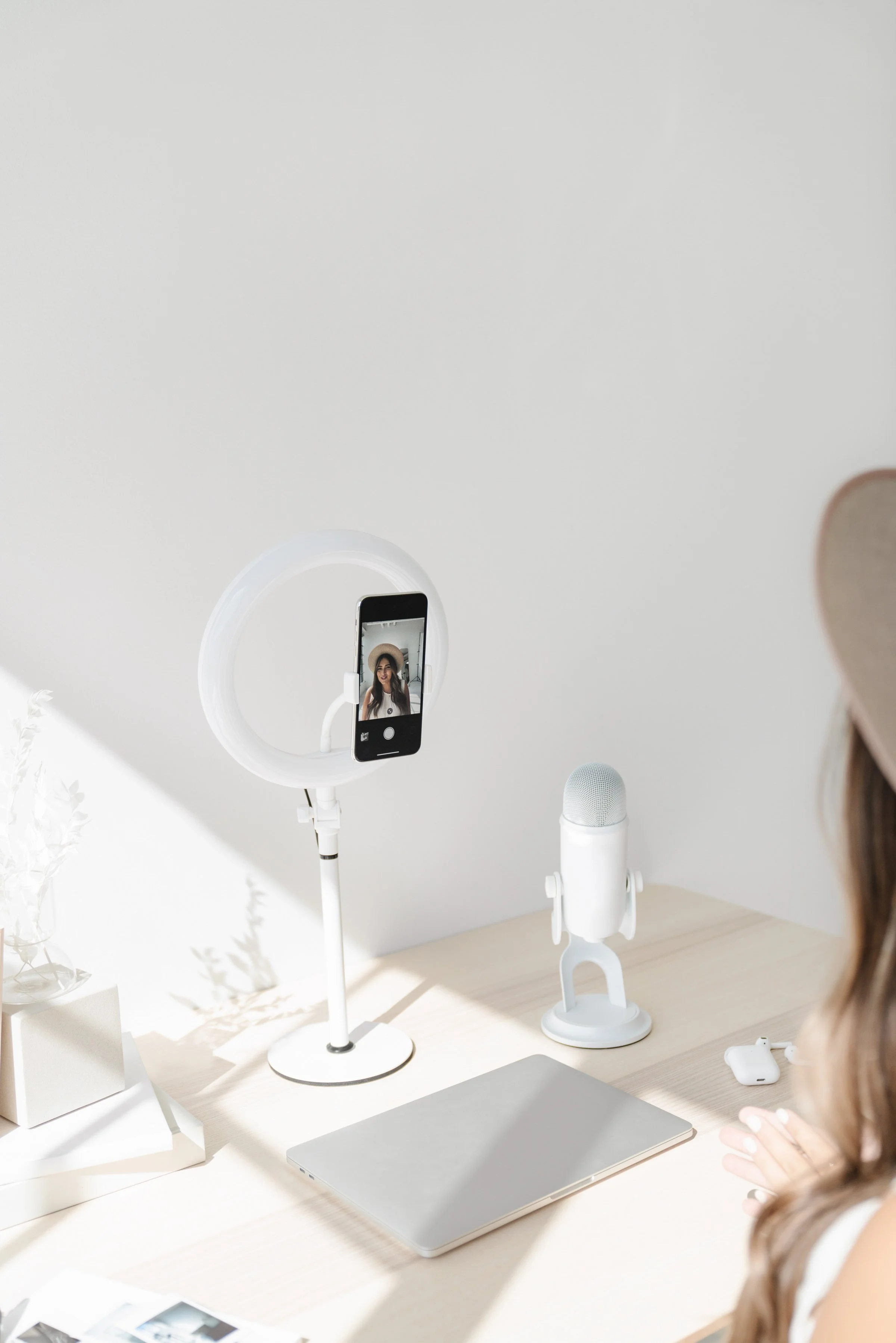 A woman sitting at a desk with a smartphone mounted on a ring light, a webcam, a closed laptop, AirPods, and some papers, all in a minimalist, white setting.