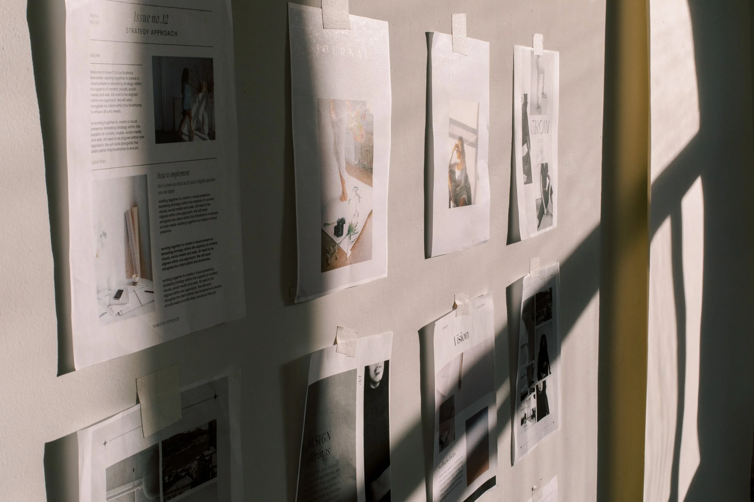 A beige wall with various papers and photographs taped to it, some are bilingual and others are in English. Shadows cast across the papers, indicating sunlight.