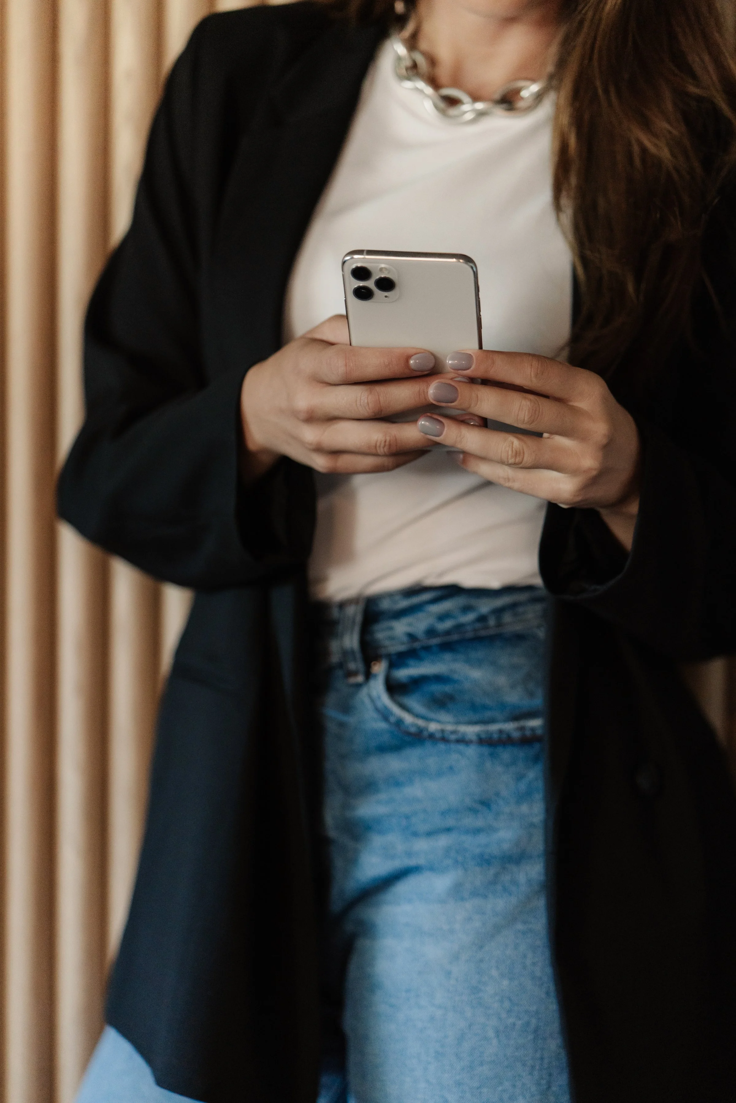 Woman holding a smartphone, wearing a black blazer, white top, blue jeans, and a chunky silver necklace.