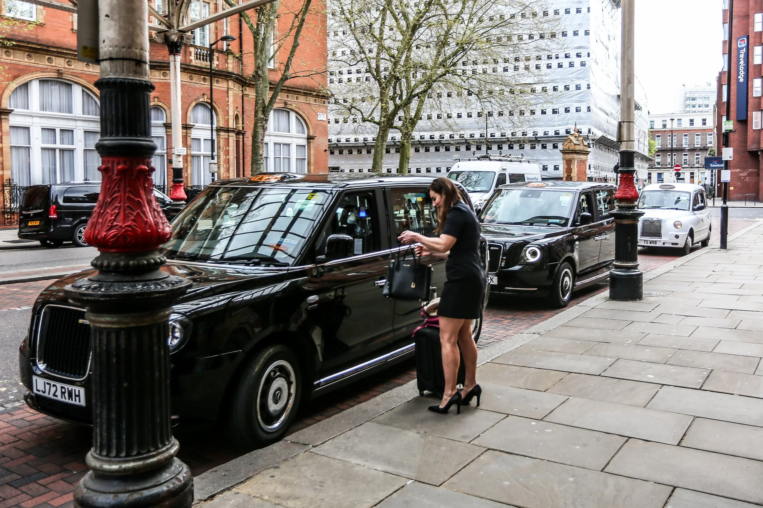 A woman in black dress and high heels stands on the sidewalk, holding a black handbag and pulling a small suitcase, next to a parked black taxi cab. Behind her are several other taxis parked along the street. The background shows a city street with old and modern buildings, some wrapped in construction material, and a large tree.