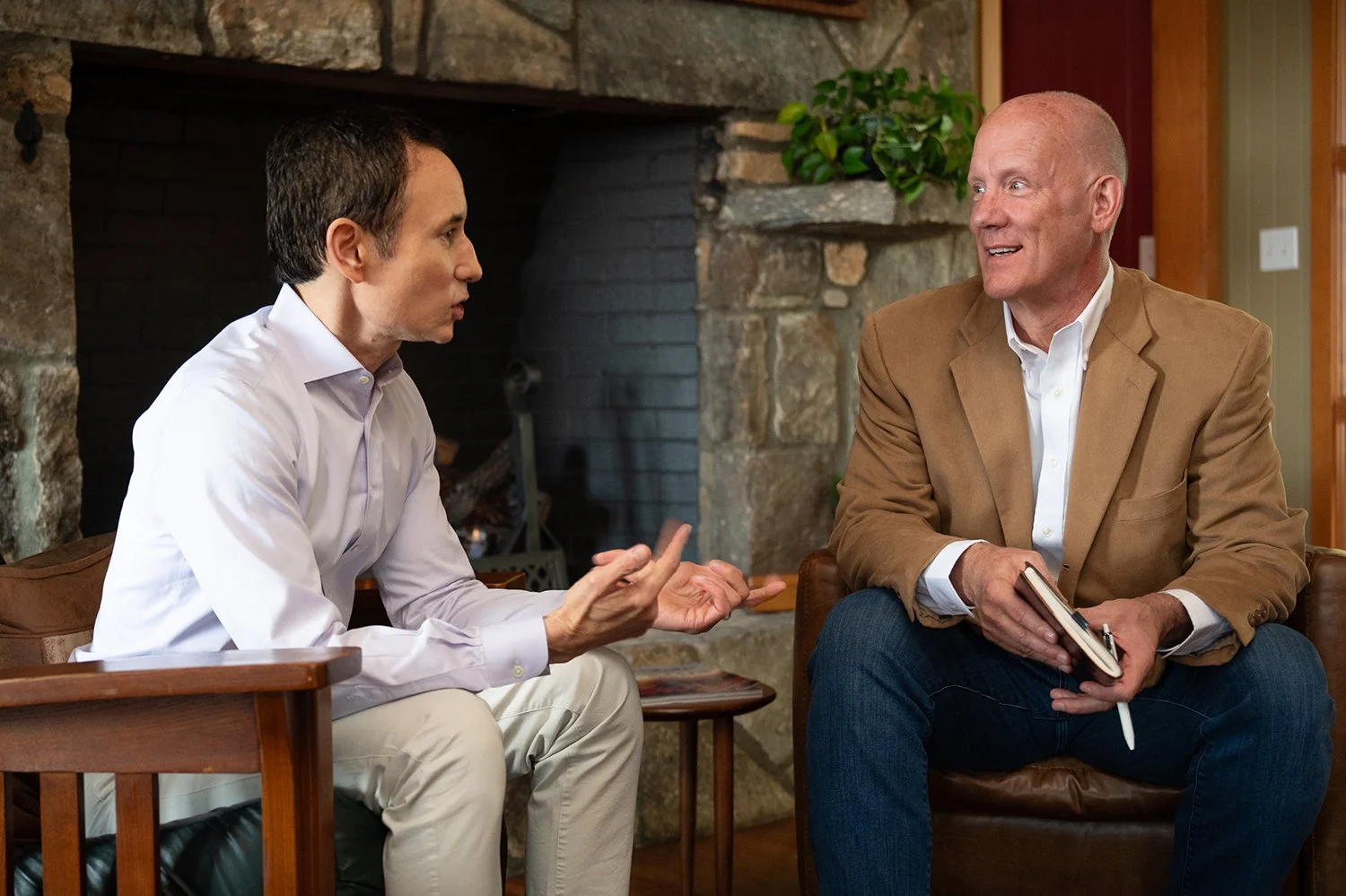 Two men engaged in conversation, sitting inside a room with a stone fireplace and wooden furniture.