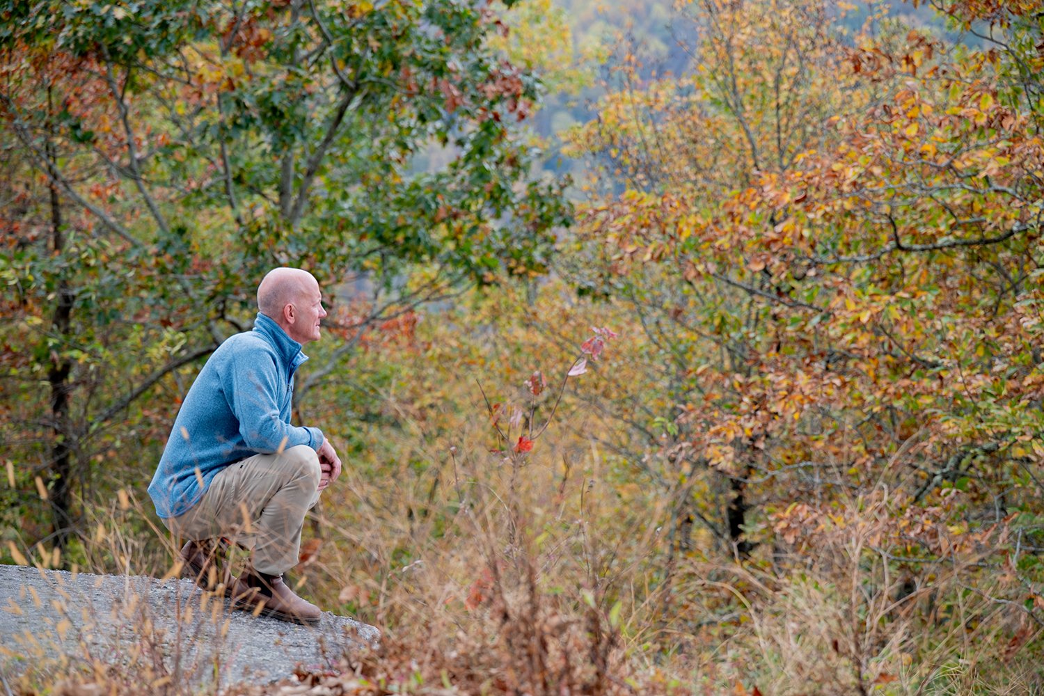 A man with a bald head, wearing a light blue fleece jacket, beige pants, and brown shoes, is squatting on a large rock in a forest with autumn foliage. He appears to be looking into the distance among trees with leaves in shades of orange, yellow, and green.