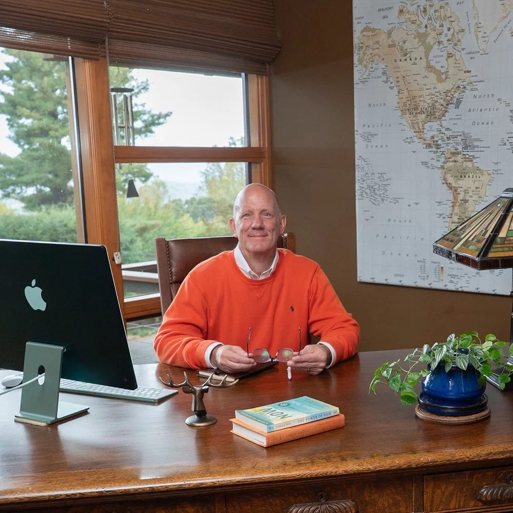 A man in an orange sweater sitting at a wooden desk in a home office, holding glasses, with a globe map on the wall behind him, a computer on the desk, and a window showing green trees outside.