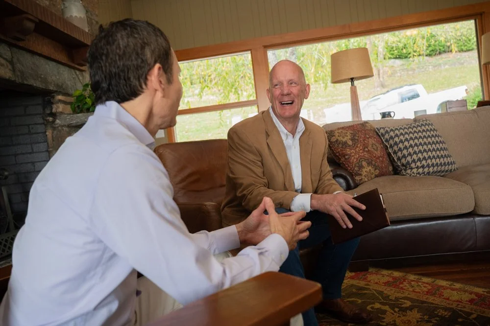 Man in white shirt and man in tan jacket having a conversation in a cozy living room with natural light, patterned pillows, and a fireplace.