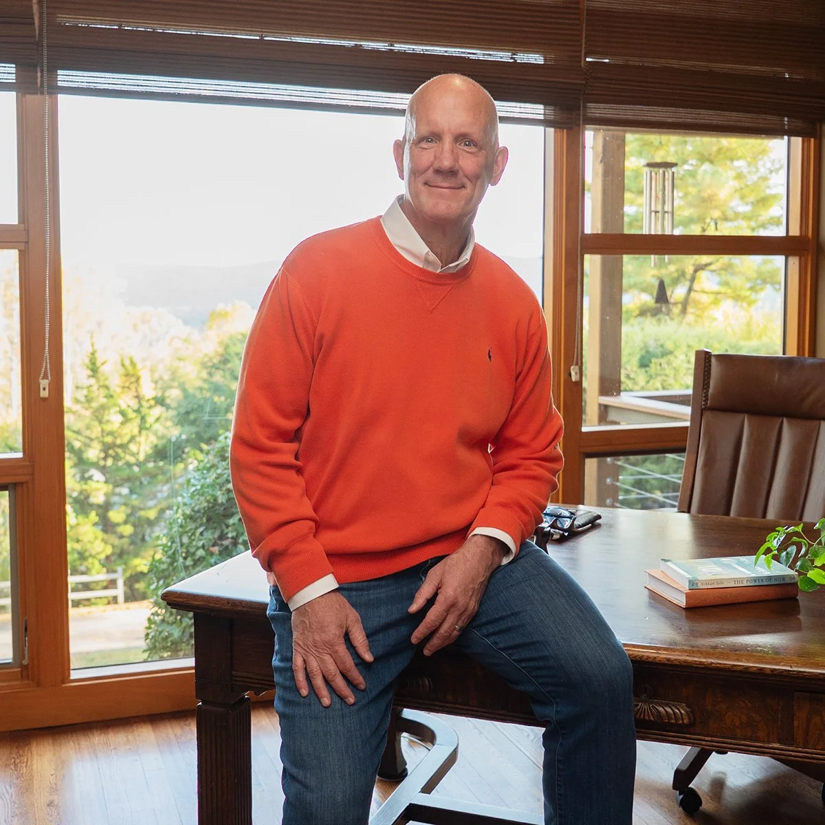 A man sitting on a wooden table in a room with large windows showing a view of trees outside, wearing an orange sweater and blue jeans.