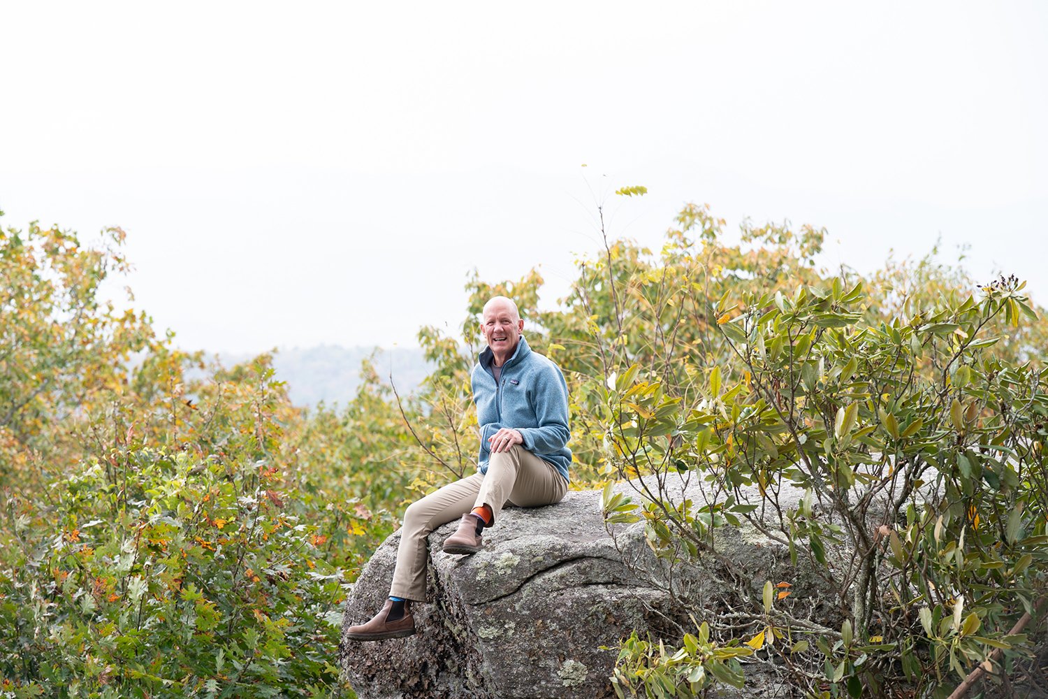 A smiling man in a blue fleece and beige pants sitting on a large rock among green shrubbery and trees with fall foliage.