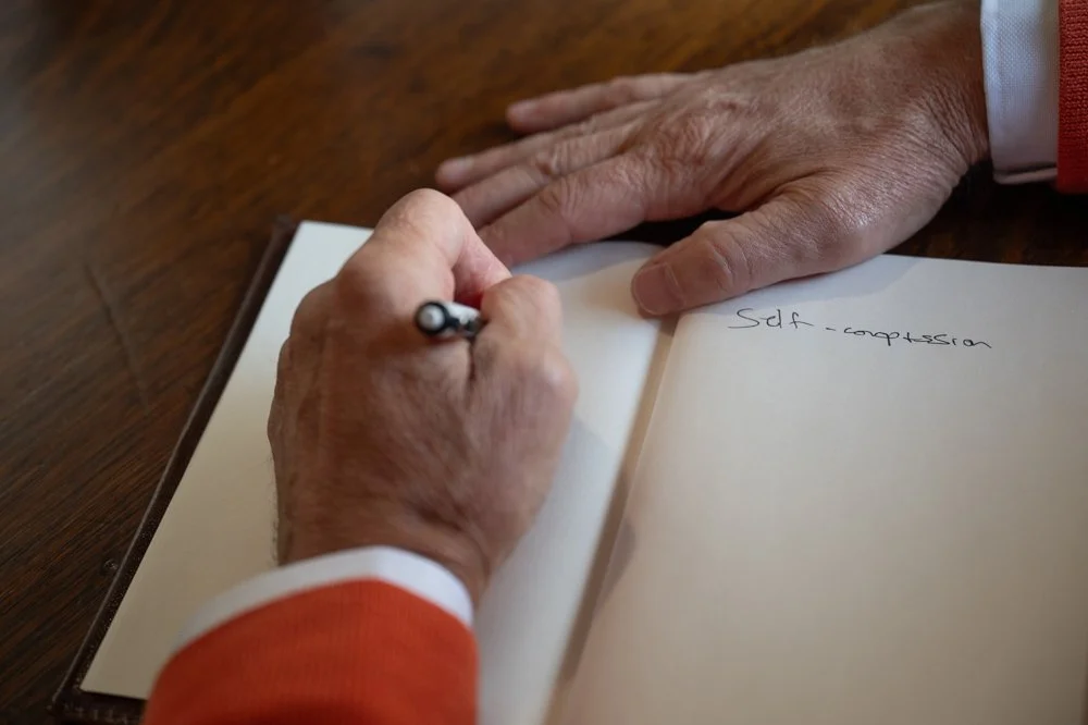A person is writing in a notebook on a wooden table, wearing an orange sleeve, with the words 'Self-compression' partially visible on the right page.