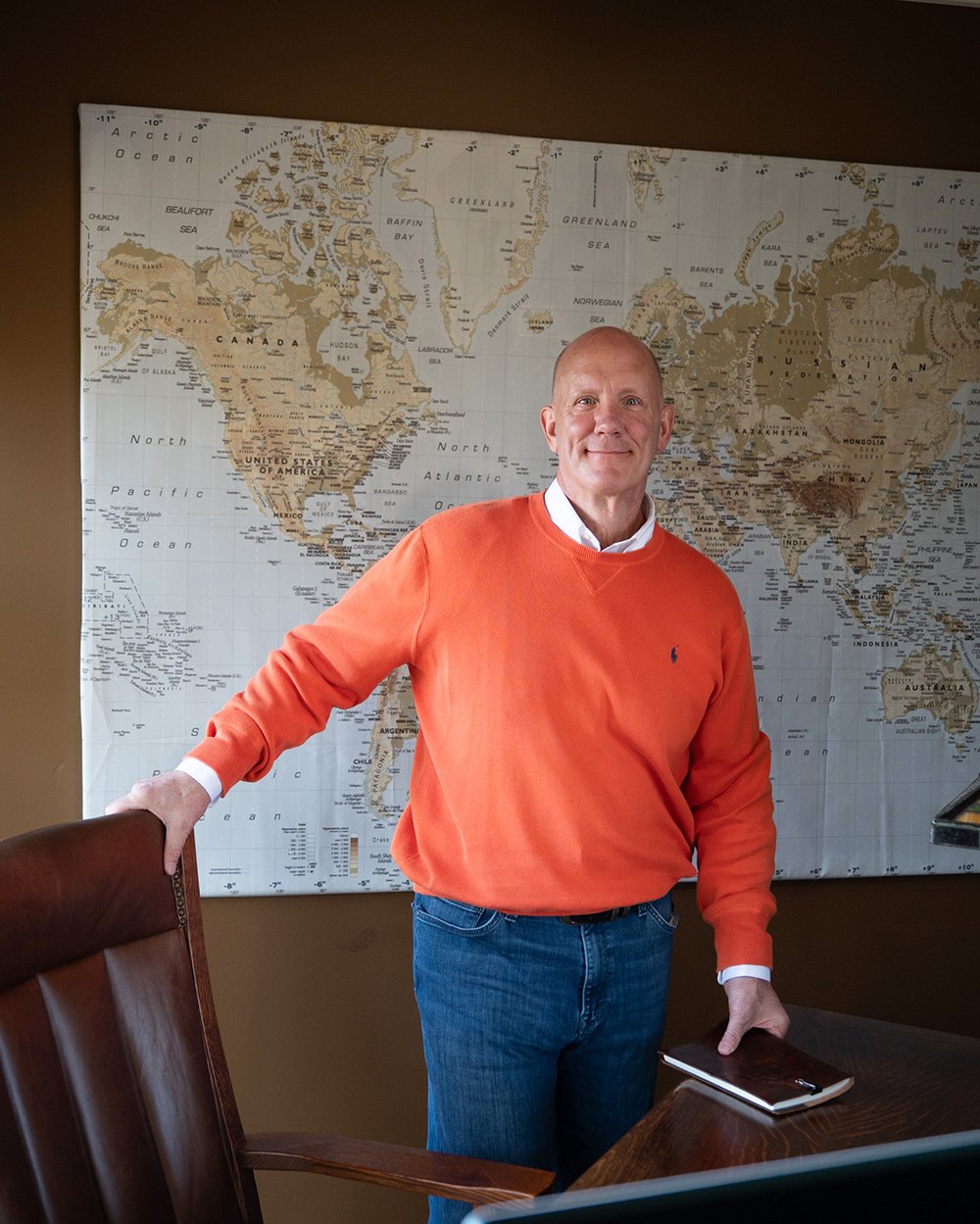 A man stands in front of a large world map, wearing an orange sweater, holding a laptop, and smiling.