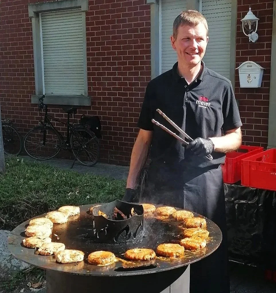 Ein Mann beim Grillen mit Tongs vor einem Grill, auf dem er Fleischstücke zubereitet. Im Hintergrund ein Haus mit rotem Backstein und Fahrrädern.