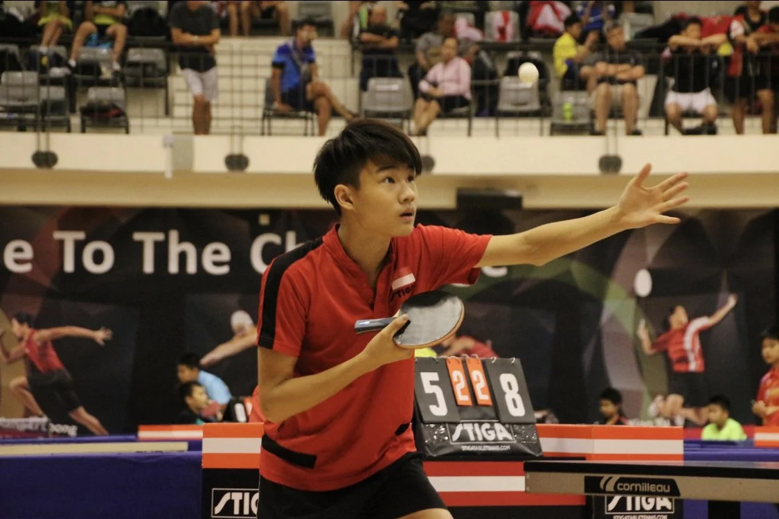 A young man playing table tennis indoors, wearing a red shirt, and preparing to hit the ball with a paddle. In the background, there are spectators seated, watching the game.  He is called Nicholas and represented Singapore for overseas competitions.
