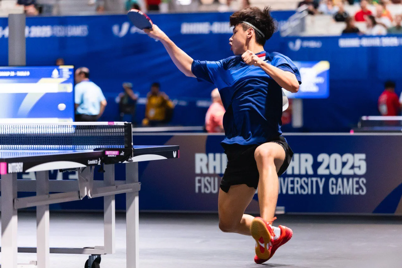 A young male table tennis player in a blue shirt and black shorts is in action, a match at the 2025 RHMR FISU University Games.  He is called Jayden and represented Singapore for overseas competition