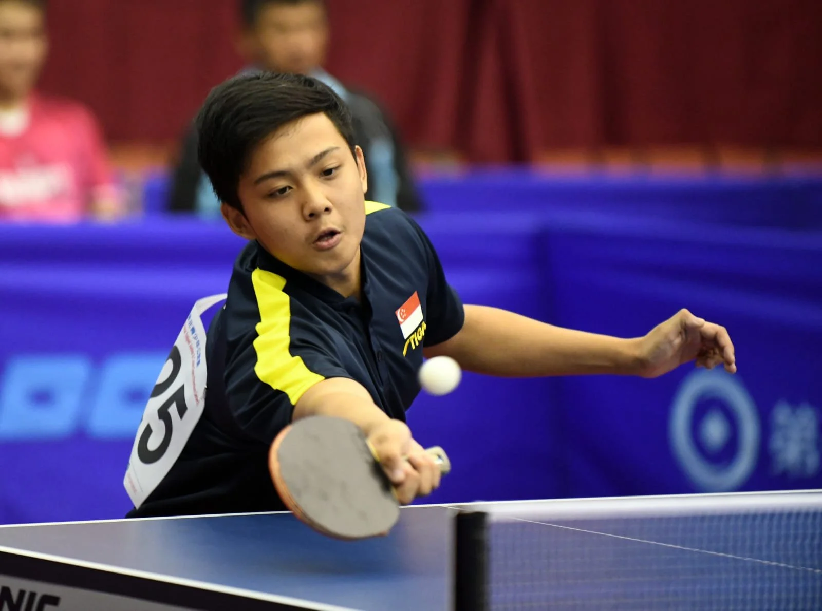 Young male table tennis player in a dark sports shirt with yellow stripes, practicing a forehand shot during a match.  He is called Andy and represented Singapore for overseas competitions.